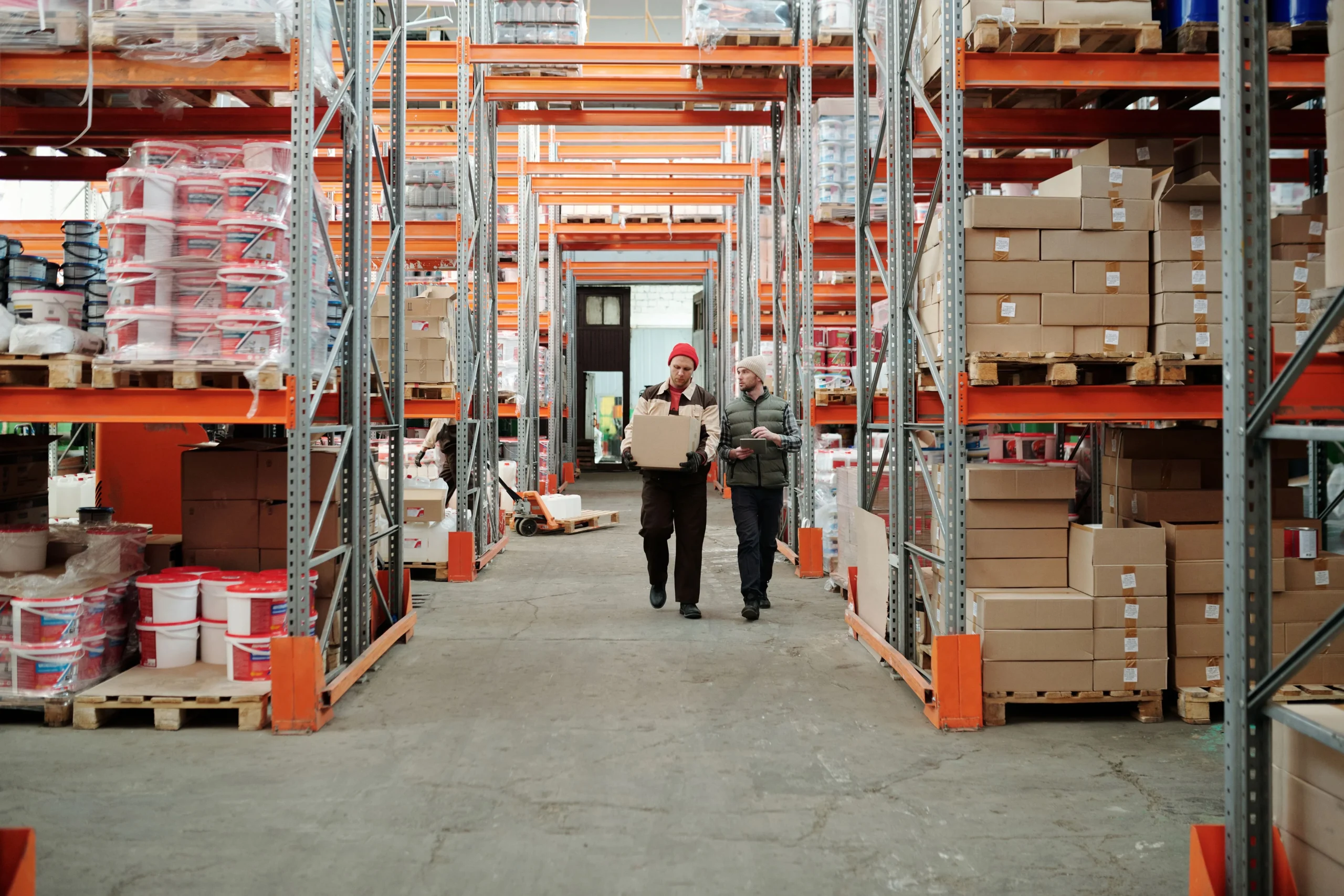 How to choose a UK e-commerce fulfilment partner 7 Two warehouse workers walking down an aisle of orange racking, one carrying a cardboard box and the other holding a tablet