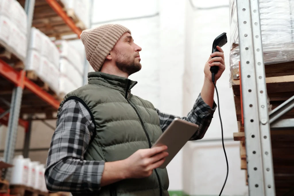 Warehouse worker using a handheld barcode scanner on a wrapped pallet while holding a tablet